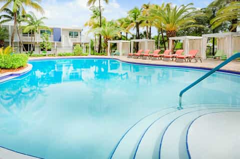 Curved resort pool with palm trees, red loungers, white pergolas and buildings