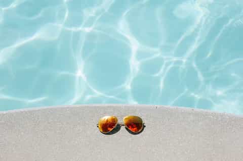 Pair of rose-gold aviator sunglasses on sand with turquoise pool water background