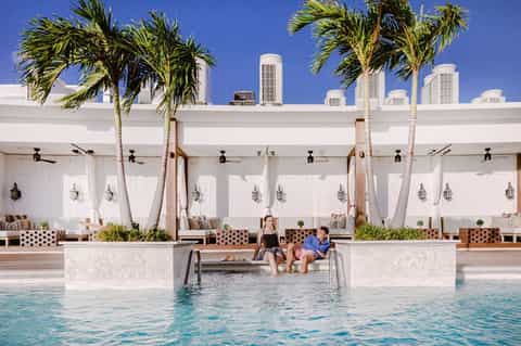 Resort pool area with white cabanas, palm trees, lounging guests, and modern architecture