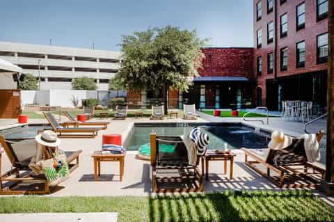 Urban resort courtyard with two pools, wooden lounge chairs, red accents, and brick building backdrop