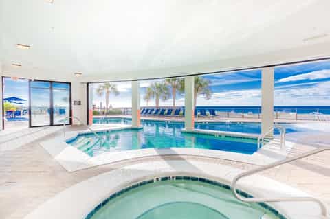 Indoor pool area with spa tub, overlooking ocean, palm trees and beach through large windows