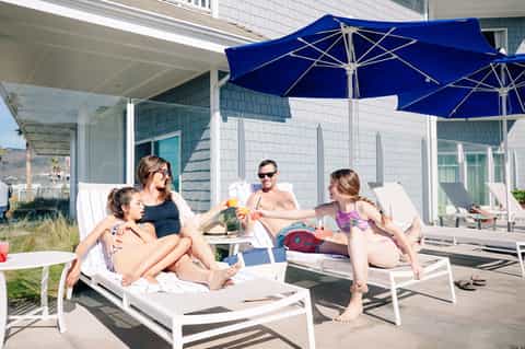 Family relaxing on lounge chairs under blue umbrella on beachfront patio with white building backdrop