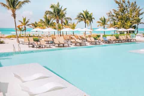 Beachfront resort pool with turquoise water, palm trees, white umbrellas, and lounge chairs