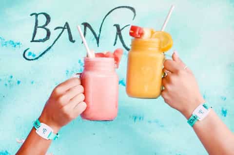 Two colorful tropical drinks with fruit garnish, festival wristbands, and 'BANK' text on turquoise background