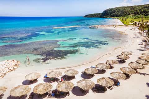 Tropical beach with turquoise water, white sand, palm trees, and thatched umbrellas lining the shore