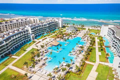 Aerial view of beachfront resort with multiple pools, palm trees, blue ocean, white buildings, sunbeds