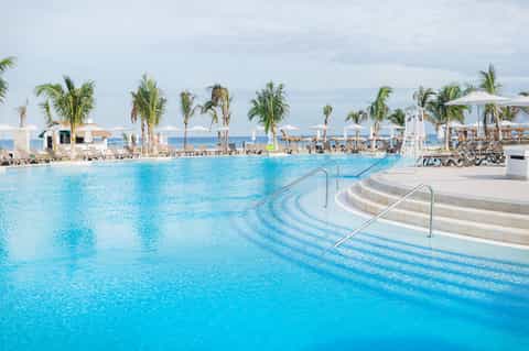 Resort-style pool with palm trees, lounge chairs, and beach in background