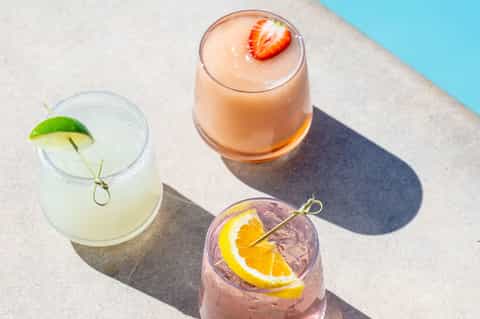 Three colorful cocktails with fruit garnishes on poolside concrete