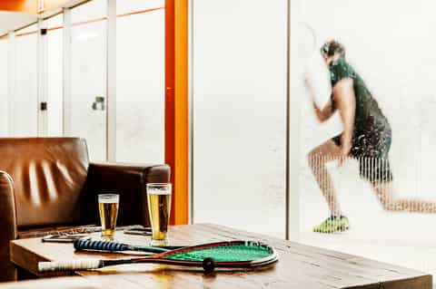 Indoor lounge with leather seating, tennis rackets, beers on wooden table, and squash player visible through glass