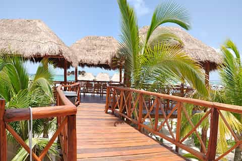 Wooden boardwalk with palm trees leading to thatched-roof beach cabanas overlooking turquoise ocean