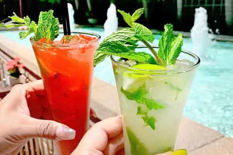 Two colorful tropical cocktails with mint garnish held by pool with turquoise water and resort lounge chairs