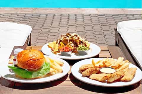 Poolside food service with burger, fried chicken strips, nachos, and condiments on table by turquoise pool