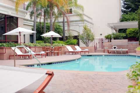 Resort pool with lounge chairs, white umbrellas, palm trees, and modern building architecture