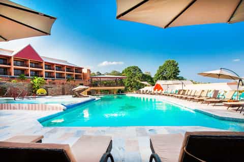Resort pool with turquoise water, sun umbrellas, lounge chairs, and red-brick building surrounded by landscaping