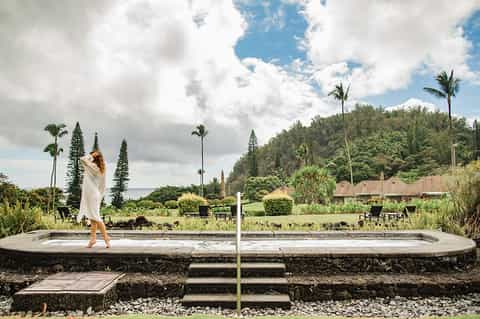 Woman in white wrap posing on stone platform near resort with palm trees, manicured gardens, and tropical vegetation