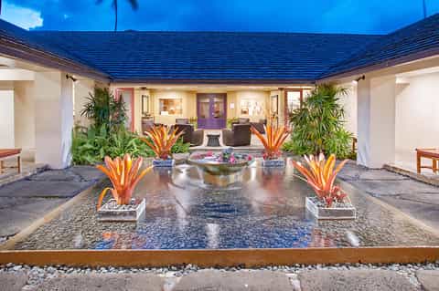 Tropical courtyard with central water feature, flowering plants, covered seating areas, and blue roof accent