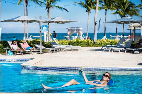 Woman floating on blue raft in pool with beach umbrellas, palm trees, and ocean view