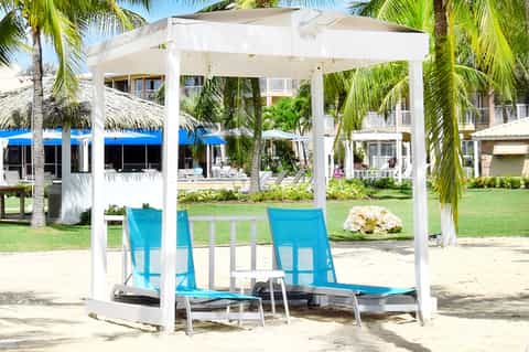 Sandy beach seating area with white pergola, blue lounge chairs, and palm trees near beachfront resort