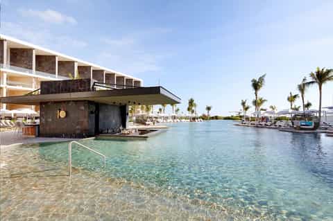 Modern resort pool with turquoise water, bar pavilion, palm trees, and lounge seating