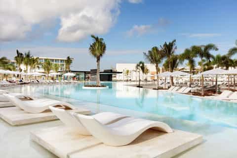 Resort pool area with curved loungers, palm trees, and white umbrellas under clear sky