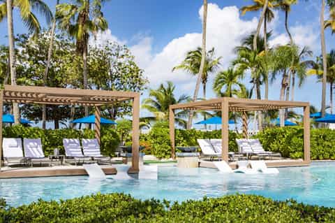 Luxurious pool area with wooden pergolas, white loungers, blue umbrellas, palm trees, and lush tropical landscaping