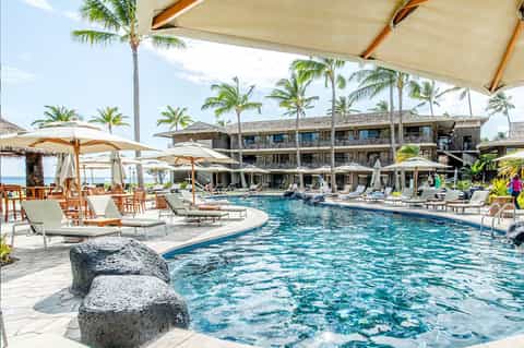 Beachfront resort pool with rock features, loungers, umbrellas, and multi-story stone building