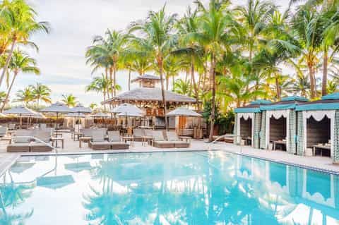 Luxurious resort pool surrounded by palm trees, lounge chairs with umbrellas, cabanas, and tropical architecture