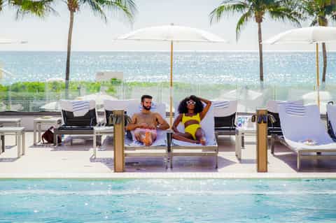 Beachfront resort pool with guests lounging, white umbrellas, ocean view, and palm trees