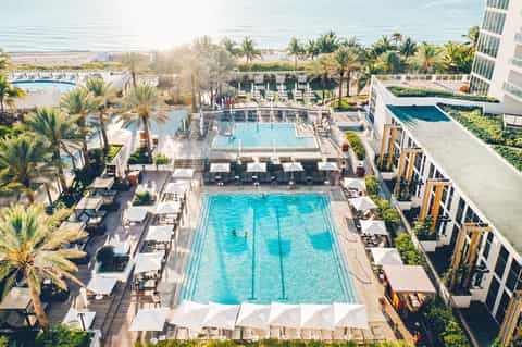 Aerial view of beachfront resort pool complex surrounded by palm trees, loungers, and modern hotel building
