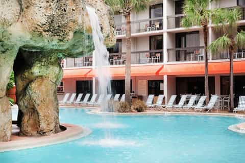 Resort pool with waterfall feature, palm trees, and multi-story coral-colored building