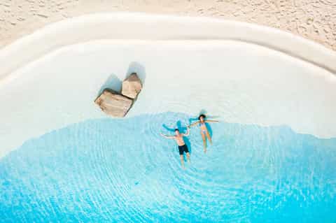 Aerial view of guests swimming in a bright blue pool shaped like a map on white sand