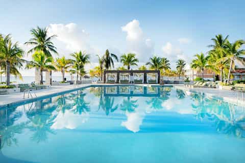 Large resort pool reflecting clouds and sky, lined with palm trees and beachfront structures
