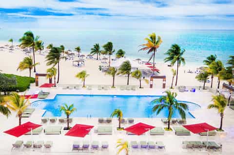Aerial view of resort pool with red umbrellas, beach, and turquoise ocean with palm trees