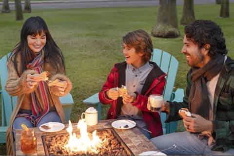 Three people enjoying s'mores around a fire pit table on a grassy lawn at dusk