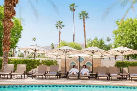 Desert poolside with lounge chairs, tan umbrellas, palm trees, mountains, and two guests relaxing
