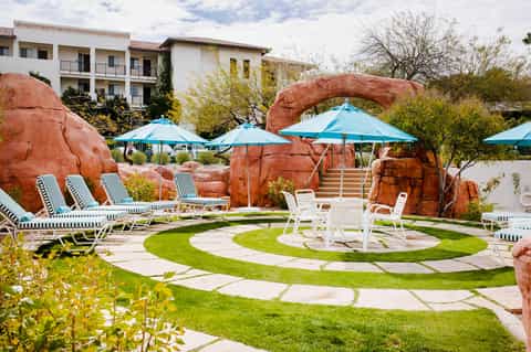 Desert-themed resort courtyard with red rock formations, blue umbrellas, lounge chairs, and manicured lawn
