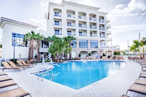 Waterfront hotel building with blue swimming pool, white lounge chairs, and palm tree landscaping