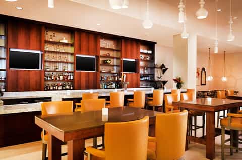 Modern bar and lounge with orange chairs, wood tables, red wood paneling, and pendant lighting