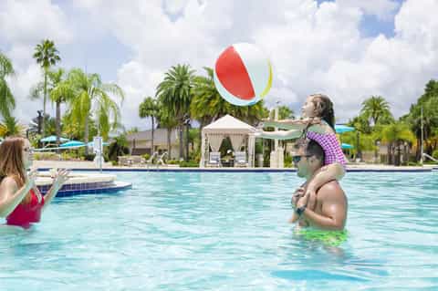 Family playing with beach ball in outdoor pool surrounded by palm trees and white cabanas