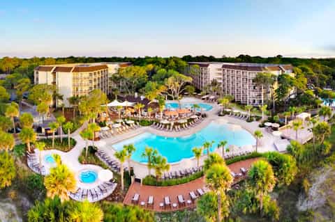 Aerial view of resort with multiple pools, palm trees, and multi-story hotel buildings surrounded by forested landscape
