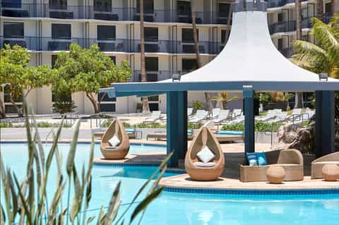 Luxury resort pool area with woven pod lounge chairs, white pergola canopy, palm trees, and modern building.
