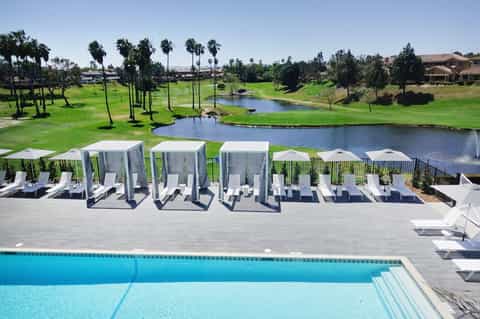 Resort pool deck with white cabanas overlooking a golf course and lake under clear blue sky