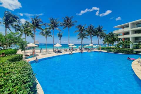 Beachfront resort pool with ocean view, palm trees, white umbrellas, and sandy beach beyond