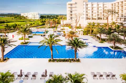 Aerial view of resort with multiple blue pools, white buildings, palm trees, and green surroundings