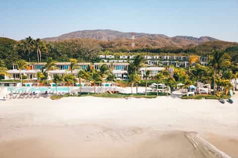 Beachfront resort with white buildings, pools, and palm trees backed by mountains