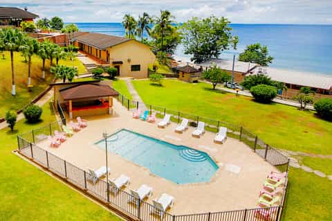 Aerial view of beachfront resort pool, white lounge chairs, palm trees, and ocean view with grassy lawn