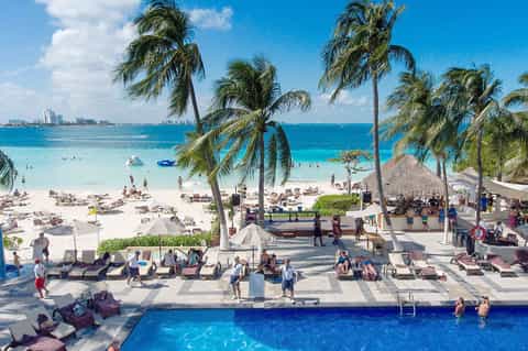 Tropical beach resort with ocean view, palm trees, pool, and crowd of sunbathers enjoying the sandy shore