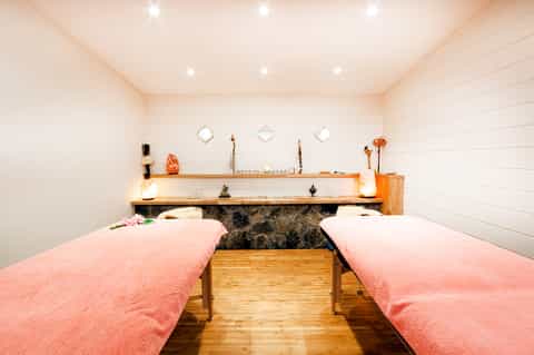 Spa treatment room with two pink massage beds, wooden shelving, and calming neutral décor with ambient lighting