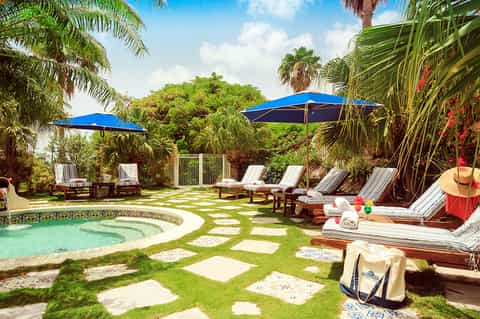 Tropical resort courtyard with spa pool, lounge chairs, blue umbrellas, palm trees, and lush landscaping