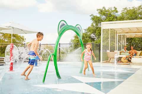 Splash pad with children playing under water fountains and green arch feature, shaded seating area nearby